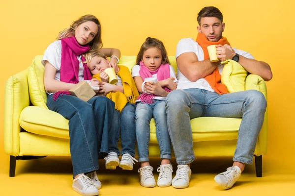 sick family with kids in scarves holding cups with hot drinks while sitting on sofa on yellow