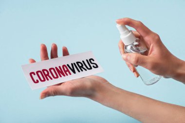 Kırpılmış bir görüntü, elinde sprey şişesiyle kartın yanında Coronavirus harfleriyle mavi renkli el dezenfektanı tutan kadın. 