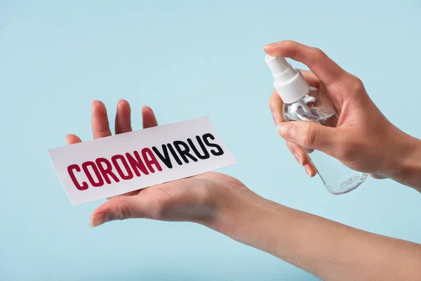 cropped view of woman holding spray bottle with hand sanitizer near card with coronavirus lettering isolated on blue