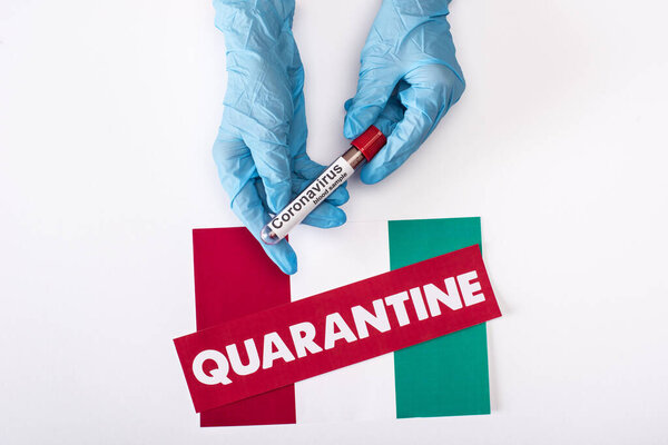 top view of doctor in latex gloves holding test tube with blood sample near quarantine lettering on italian flag isolated on white 