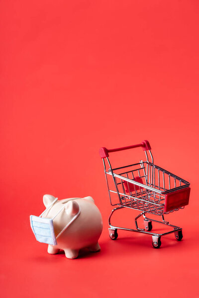 piggy bank in small medical mask near toy shopping trolley on red 