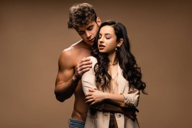Üstsüz adam bej ceketli, yarı çıplak seksi kız arkadaşına sarılıyor.