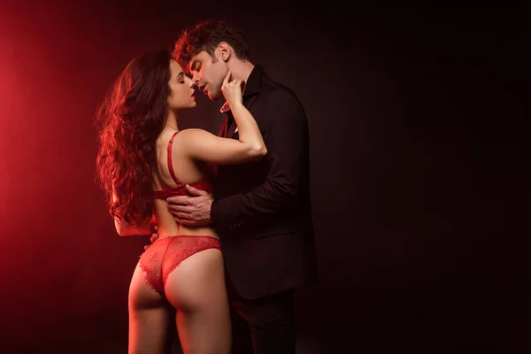 スーツ姿の男抱擁とキスへ行きますセクシーガールフレンドで赤ランジェリーで黒とともに赤ライト — ストック写真