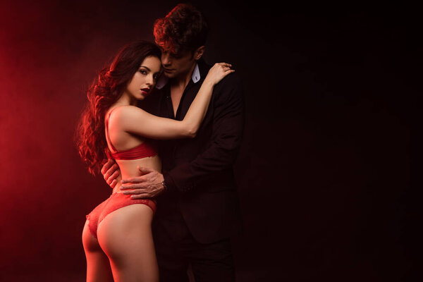 man in suit hugging sexy girlfriend in red lingerie on black with red light