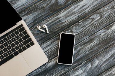 Diz üstü bilgisayar, kulaklık ve ahşap yüzey üzerinde boş ekranlı akıllı telefon.