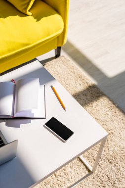 Kanepeli oturma odası, dizüstü bilgisayarlı masa, akıllı telefon ve gün ışığında not defteri.