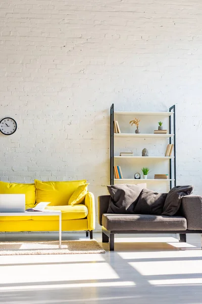 living room with yellow sofa, grey armchair, shelf and laptop in sunlight