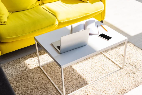 living room with yellow sofa and table with laptop, smartphone and notepad in sunlight