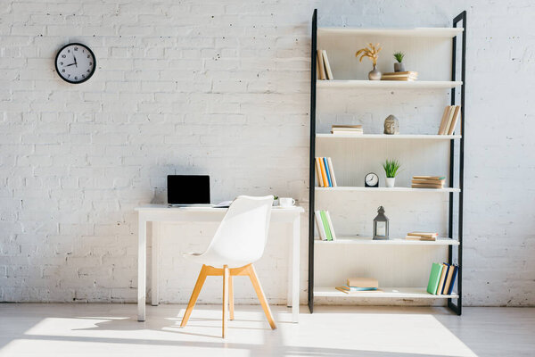 home office with book shelf, clock, chair and laptop on table in sunlight