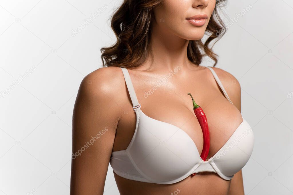 cropped view of sexy girl with red hot chili pepper between big breasts isolated on white