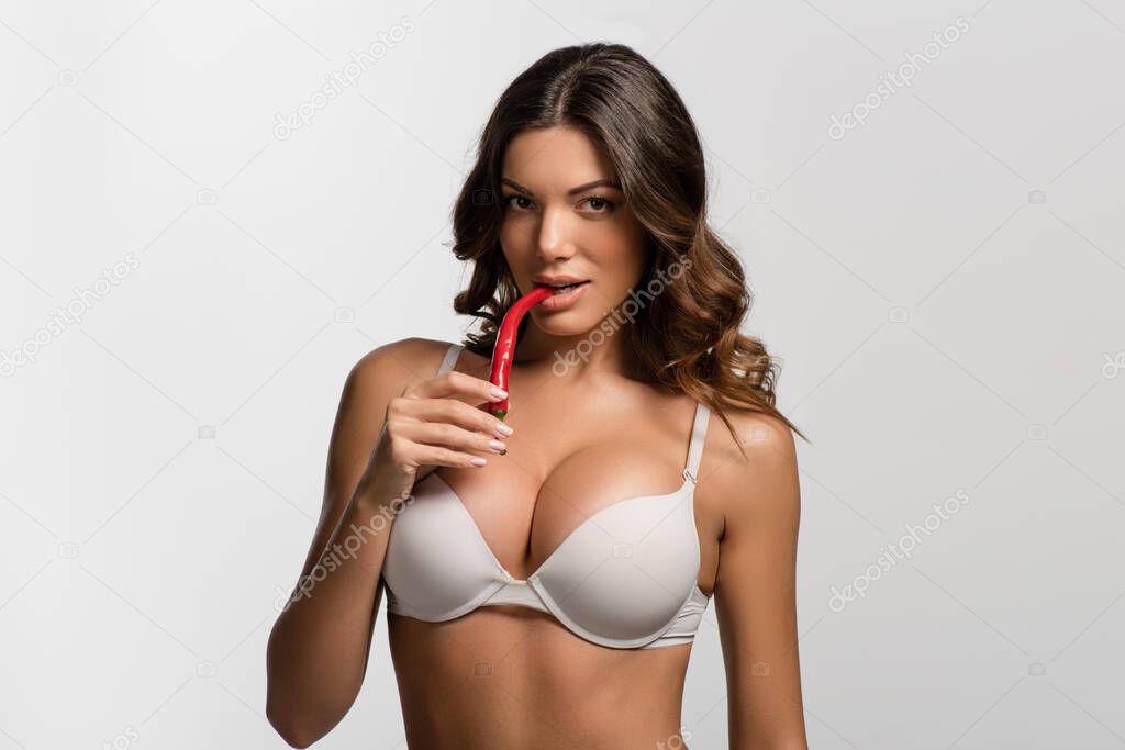 flirty, seductive girl with big breasts holding red hot chili pepper near lips while looking a camera isolated on white