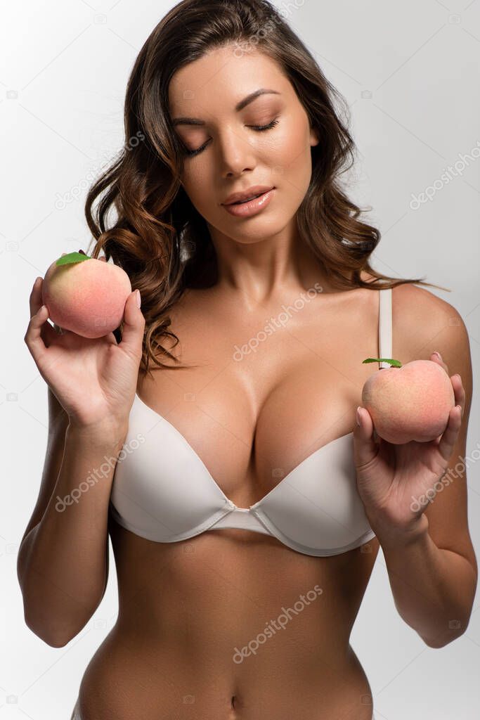 sexy, pensive girl with big breasts and closed eyes holding ripe peaches isolated on white