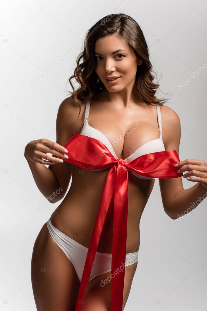 sexy, flirty girl with big breasts touching satin bow on bra while looking at camera isolated on white