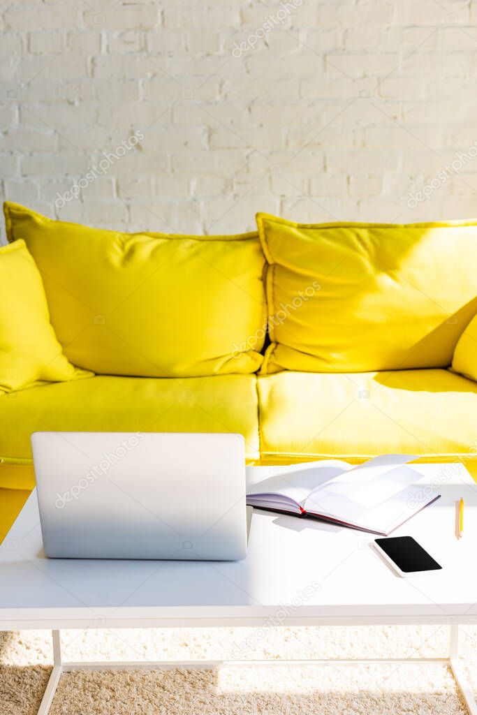 living room with yellow sofa and table with laptop, smartphone and notepad in sunlight