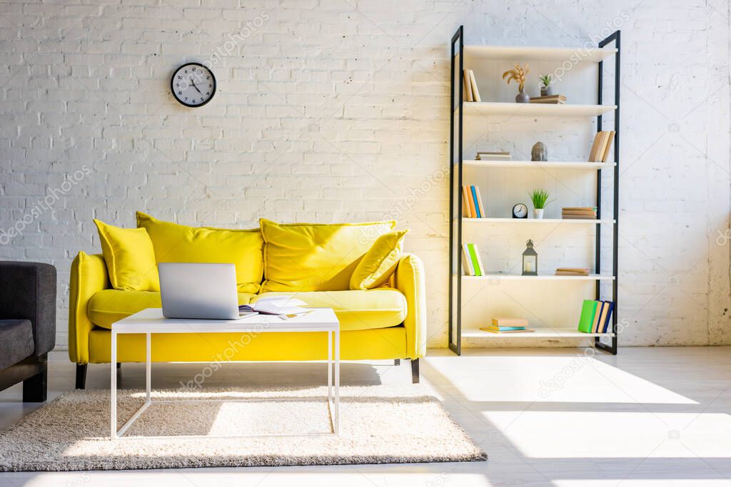 living room with yellow sofa, shelf and table with laptop in sunlight