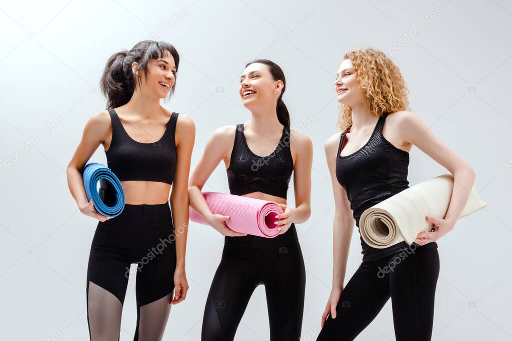 happy and multicultural women holding fitness mats on white 