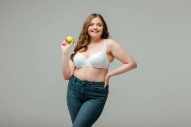 happy plus size woman in jeans and bra holding apple isolated on grey clipart