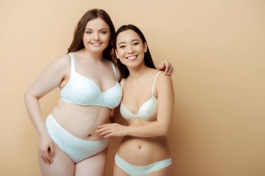 plus size woman smiling with asian girl in underwear isolated on beige, body positive concept  clipart