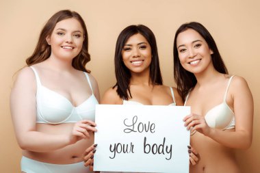 cheerful and overweight multicultural women in bras holding placard with love your body lettering isolated on beige, body positive concept  clipart