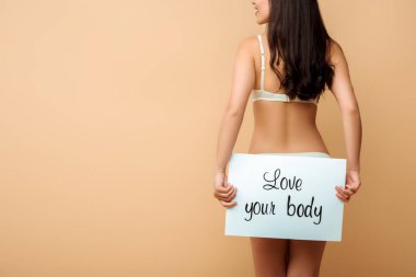 cropped view of happy girl holding placard with love your body lettering isolated on beige  clipart