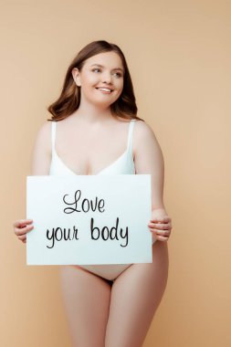 cheerful plus size woman holding placard with love your body lettering isolated on beige, body positive concept  clipart