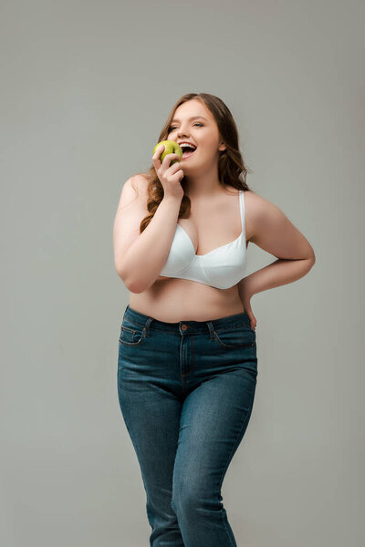 веселая плюс размер женщина в джинсах и бюстгальтер едят яблоко изолированы на серый
