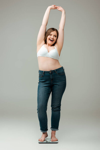 happy plus size woman in jeans and bra standing with hands above head on scales and smiling on grey