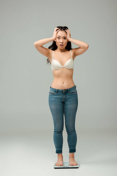 шокированная азиатка в джинсах и лифчике, стоящая на весах и трогательная голова на серой
