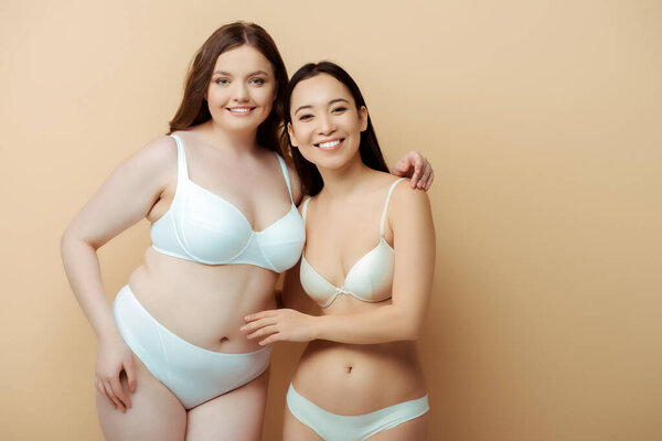 plus size woman smiling with asian girl in underwear isolated on beige, body positive concept 