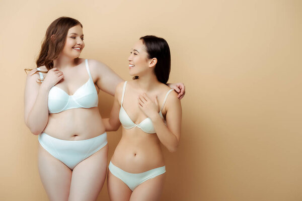 happy plus size woman standing with asian girl in underwear isolated on beige, body positive concept 