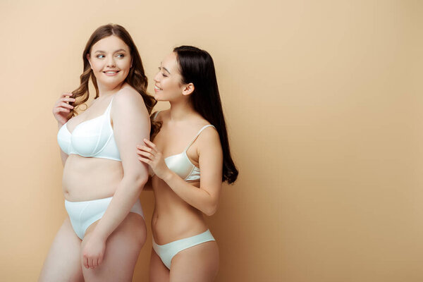 cheerful plus size woman standing with asian girl in underwear isolated on beige, body positive concept 