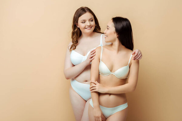 happy plus size woman hugging asian girl in underwear isolated on beige, body positive concept 