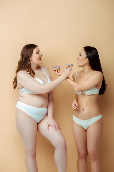 profile of happy plus size woman and asian girl in underwear holding doughnuts isolated on beige, body positive concept 