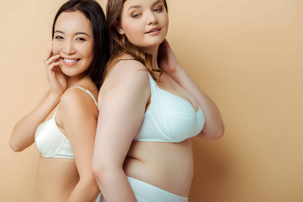 plus size woman and happy asian girl in underwear standing on beige, body positive concept 