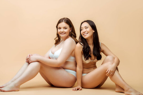 cheerful overweight woman and happy asian girl in underwear sitting on beige, body positive concept 