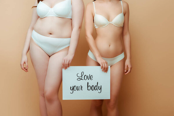 cropped view of plus size women in underwear holding placard with love your body lettering isolated on beige, body positive concept 