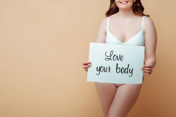 Cropped view of woman smiling and presenting placard with love your body lettering on beige background