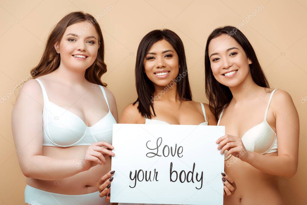 cheerful and overweight multicultural women in bras holding placard with love your body lettering isolated on beige, body positive concept 
