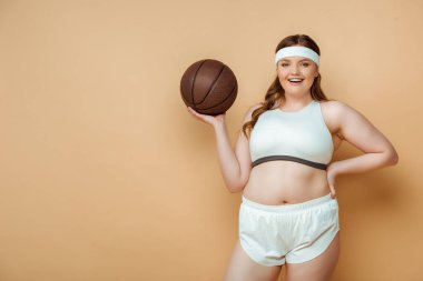 Ayrıca eli kalçasında ve topunda olan, gülümseyen ve bej renkli kameraya bakan sporcu kadın.