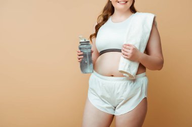 Spor şişesi ve bej rengi havluyla gülümseyen büyük beden kadın manzarası.