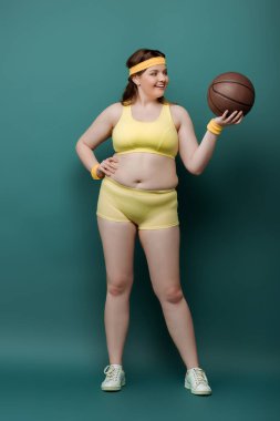 Ayrıca eli kalçasında bir sporcu kadın topa bakıyor ve yeşil arka planda gülümsüyor.