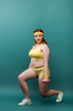 Artı, spor kadını dambıllarla hamle yapıyor ve yeşil arka plana bakıyor.