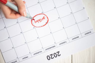 Cropped view of woman pointing with marker pen on birthday lettering in to-do calendar with 2020 inscription on wooden background clipart