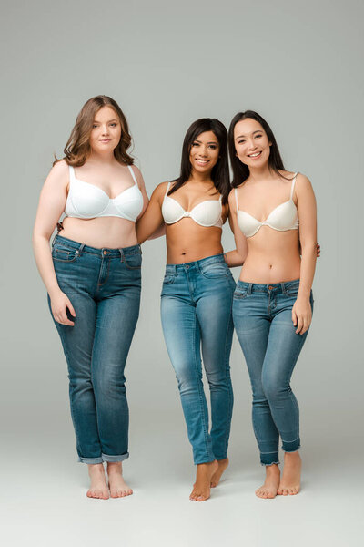 Multicultural women in bras looking at camera and smiling on grey background