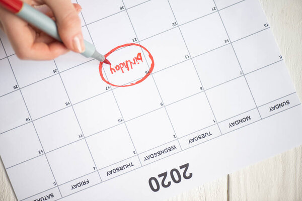 Cropped view of woman pointing with marker pen on birthday lettering in to-do calendar with 2020 inscription on wooden background