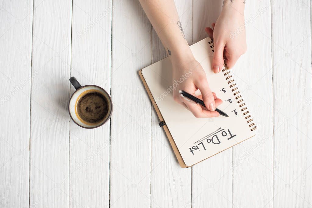 Cropped view of woman writing in notebook with to do list lettering near cup of coffee on wooden background