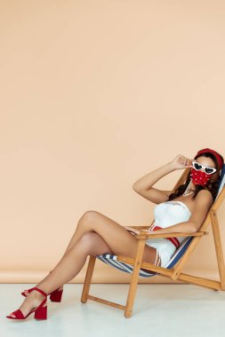 Maskeli ve mayolu bir kız bej rengi sandalyede otururken güneş gözlüklerine dokunuyor.