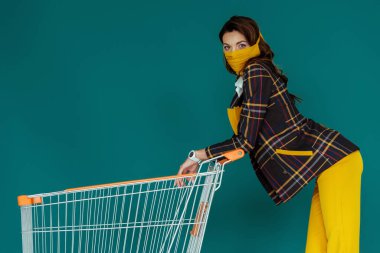 Sarı maskeli şık bir kadın elinde boş bir alışveriş arabasıyla duruyor.