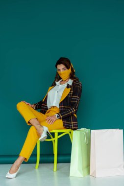Sarı maskeli, ekose ceketli şık bir kadın mavi alışveriş torbalarının yanında otururken poz veriyor.