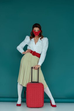 Maskeli şık bir kadın. Mavi çantanın yanında eli kalçasında duruyor. 